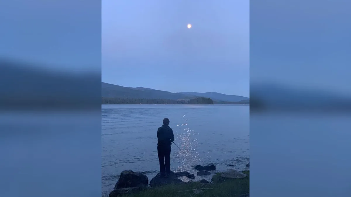 Angler mit Rute am Seeufer bei Mondaufgang in der Dämmerung